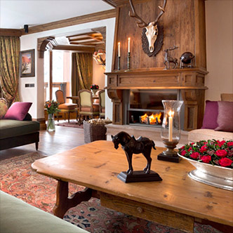 Cozy living room in Jardin Alpin apartment, Courchevel 1850: burning fireplace, antler chandelier, green armchairs, wooden interior and warm alpine decor.