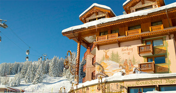 Exterior of the La Loze residence in Courchevel: traditional alpine chalet covered in snow, with deer frescoes, balconies and a passing ski lift against a backdrop of snow-covered fir trees.
