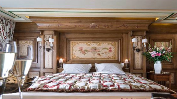 Luxury bedroom in La Loze apartment, Courchevel 1850: large bed with carved wooden headboard and fresco, champagne glasses and flower bouquet on bedside table.