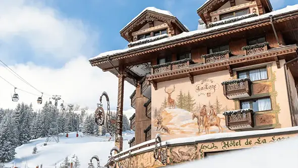 Exterior facade of Hotel de La Loze featuring traditional Savoyard wall paintings against the backdrop of a ski lift in Courchevel 1850.