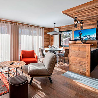Bright open living-dining room in Le Blanchot apartment, Val d&rsquo;Is&egrave;re: orange armchair, transparent chairs, panoramic windows with snowy slope view.