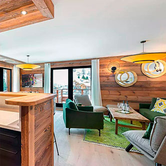 Modern living room in L&rsquo;Ourson apartment, Val d&rsquo;Is&egrave;re: green sofa and armchair, bar counter, panoramic windows with snowy mountain view and balcony.