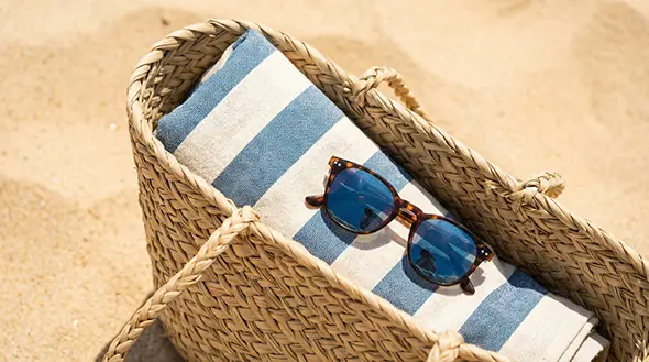 Woven beach bag with a striped towel and sunglasses on the sand.