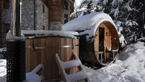 Outdoor wooden barrel sauna and hot tub in the snow by the hotel.
