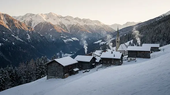Old timber hamlet and church under fresh snow at dawn with chimney smoke above the valley