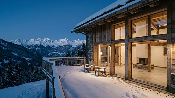 Alpine chalet terrace under fresh snow at blue hour with lit windows and mountains beyond
