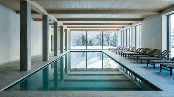Empty indoor swimming pool with stone columns and loungers facing a snow-covered forest