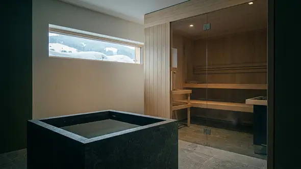 Sauna behind glass beside a dark stone cold plunge, with a snow-filled window between them