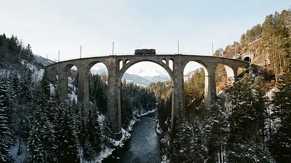 Swiss mountain train crossing a stone arch viaduct above a snow-covered forested gorge