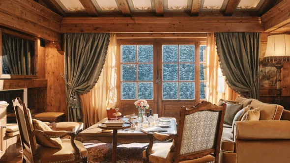 Suite living room with panelled walls, a laid table and snow-covered firs beyond the windows at Les Airelles Courchevel