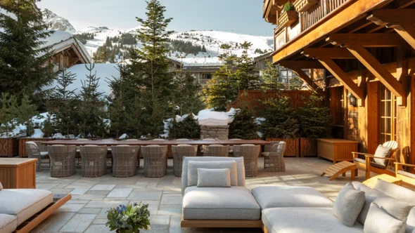 Stone terrace with low sofas, a long dining table and fir trees at Les Chalets des Airelles, Courchevel