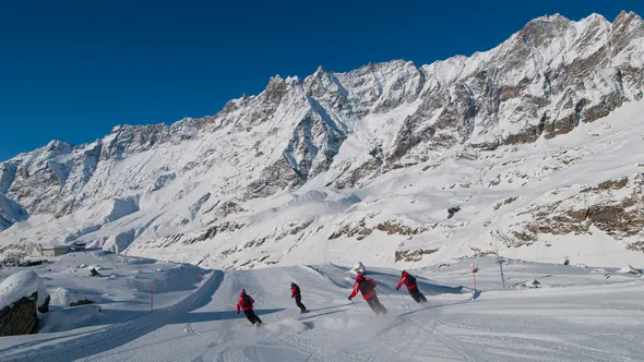 Four skiers on a wide groomed piste below a snowy ridge