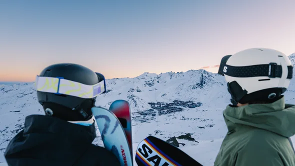 Two skiers in helmets looking down at Val Thorens from the slopes at sunset