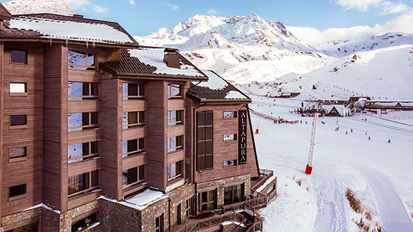 Exterior view of the 5-star Altapura hotel in Val Thorens against the snowy mountains, showcasing the modern wooden facade, direct slope-side location, and ski lift adjacent to the hotel.