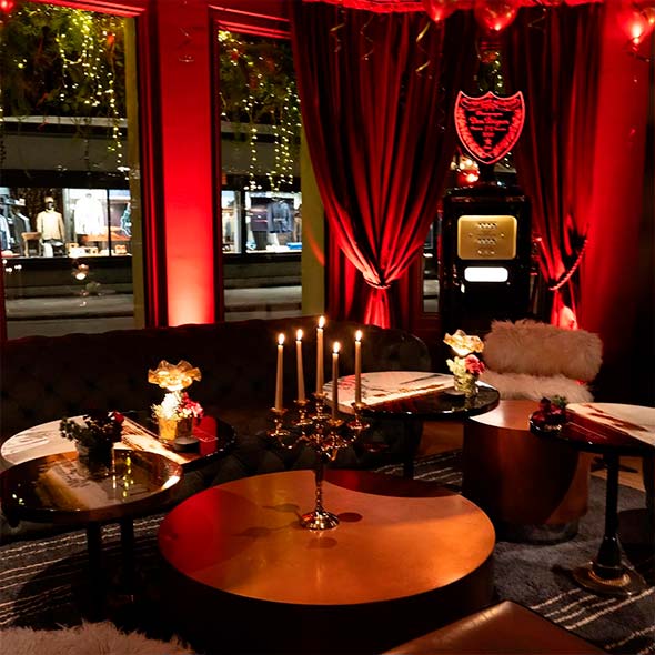 Cozy VIP corner in Balthazar St. Moritz bar with a velvet sofa, gold tables, a candle chandelier, and red curtains against windows adorned with garlands.