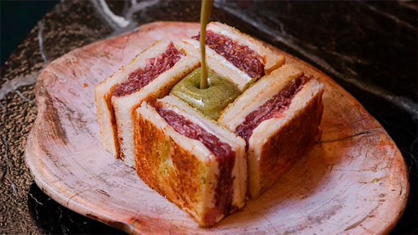 Premium Croque Sando with marbled beef (Wagyu) filling in toasted bread, being poured over with a green sauce, on a wooden plate