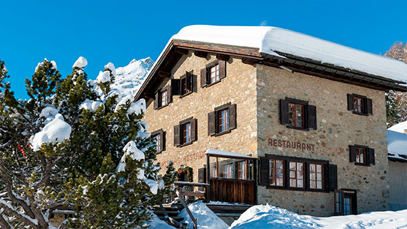 Exterior view of the traditional alpine Chasellas restaurant building made of stone with wooden shutters, situated among snow-covered trees on a sunny winter day.