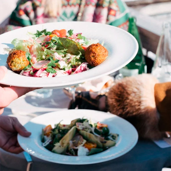 Waiter&rsquo;s hands serving a plate of green salad with meatballs (possibly falafel) at a table on the terrace.