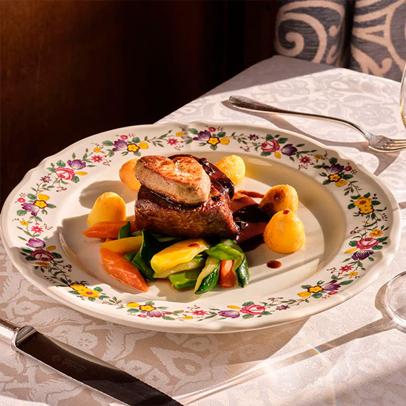 Exquisite dish of steak topped with foie gras, served with vegetables and potatoes in sauce, presented on a plate with a floral pattern.
