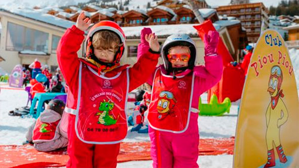 Two young child skiers in bright red suits and Piou Piou Club vests stand on the snow with hands raised, symbolizing fun and safety during their first skiing lessons in Courchevel.
