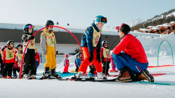 An ESF ski instructor in a red jacket assisting a young student in bright gear on a gentle, snowy slope at the Village des Enfants in Courchevel, showcasing personalized children&rsquo;s ski instruction.
