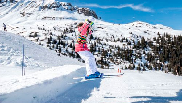 A girl in a bright ski suit performing a jump from a small snow ramp against the backdrop of snowy alpine mountains and blue sky, demonstrating freestyle skiing skills in Courchevel.