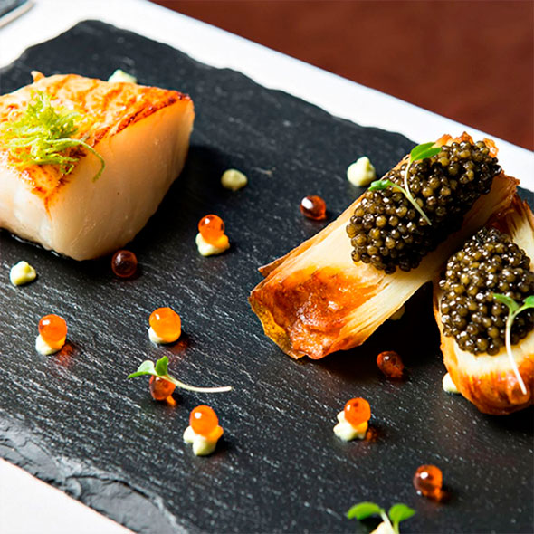 Exquisite dish of seared fish and two roasted vegetable pieces topped with black caviar, served on a black slate plate.