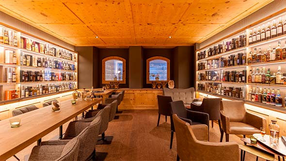 Interior of the Devil&rsquo;s Place tasting room featuring light wood paneling and ceiling, two rows of armchairs and a long table between shelves with illuminated whisky bottles.