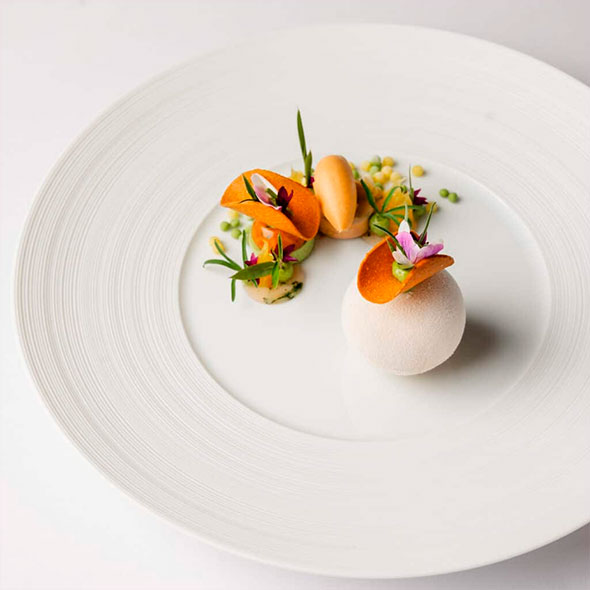 Exquisite signature dish at Ecco St. Moritz, presented as a white sphere with a crunchy orange element and delicate green sprouts on a large white plate.
