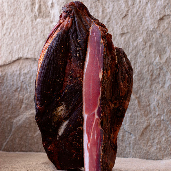 Close-up of a cut of high-quality cured or smoked meat with visible fat marbling, standing upright against a stone background.