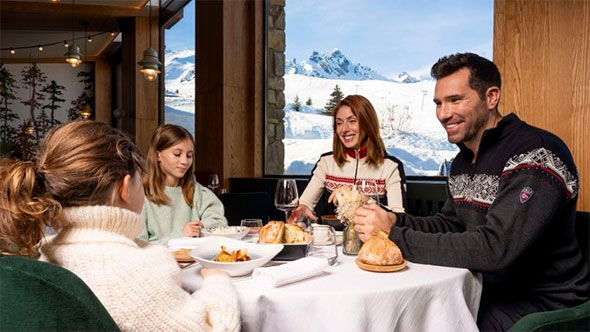 A happy family in ski sweaters dining at H&ocirc;tel Annapurna&rsquo;s restaurant, enjoying gourmet food and a panoramic view of the snowy Alps through large windows.