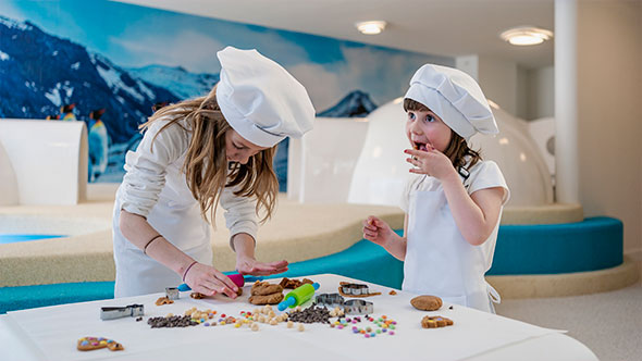 Two young girl chefs in white hats and aprons participating in a cooking masterclass (cookie baking) in the Annapurna Hotel kids club, highlighting family-friendly activities.