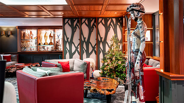 Luxurious lobby of the Annapurna Hotel with a wooden coffered ceiling and red leather sofas, decorated for Christmas with a tree and a skier mannequin in ski gear, highlighting alpine chic and art decor.