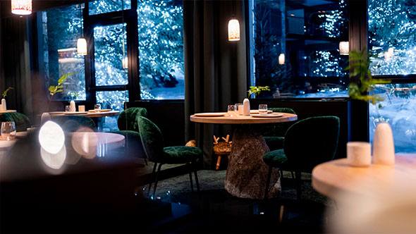 Elegant interior of La Table de l&rsquo;Annapurna restaurant featuring velvet green chairs and round tables, overlooking a snowy landscape decorated with twinkling string lights on trees, creating a festive ambiance.