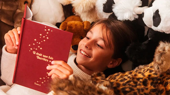 Close-up of a smiling girl lying amidst plush toys and reading a red Barrière book with gold embossing, highlighting family-friendly and cozy leisure at the hotel.