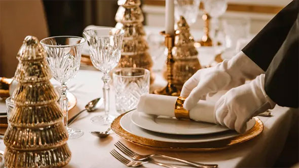 Close-up of an elegant table setting with golden Christmas tree decorations and crystal glasses, where white-gloved hands (staff) are adjusting a napkin, emphasizing the high service of the festive dinner.