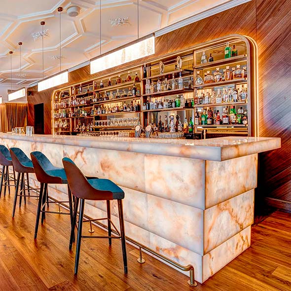 Stylish and spacious lounge bar with a high ceiling, decorative stucco, a long white stone bar counter, and a lounge area with soft seating. Ideal for day-time apr&egrave;s-ski.