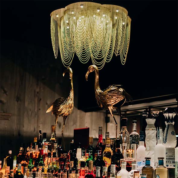 Close-up of the King&rsquo;s Social House bar counter with alcohol shelves, adorned with two golden flamingo statues and a massive tiered chandelier.