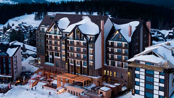 Panoramic view of the luxurious, snow-covered Viceroy Kopaonik hotel building against the backdrop of snowy mountains at sunset.