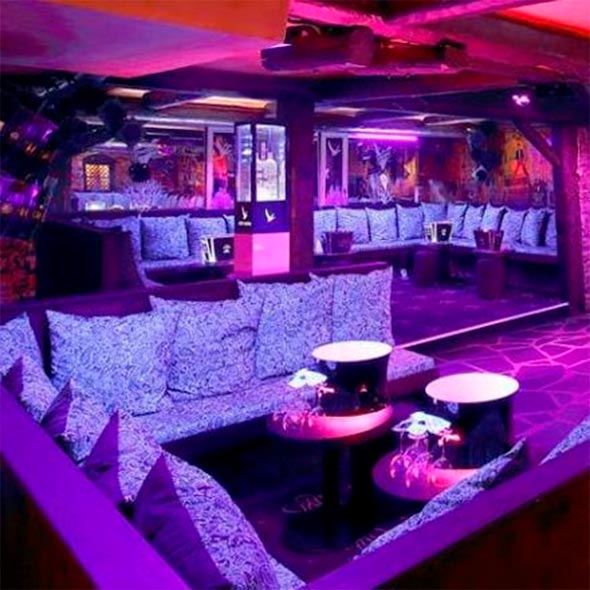 Interior of La Grange nightclub, bathed in bright purple light: VIP zones with low sofas and patterned pillows, ottoman tables, dark flooring, and wooden ceiling beams.