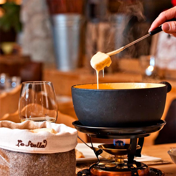 Close-up of a pot of melted cheese fondue, with a piece of bread being dipped by a fork, against a branded bread basket labeled La Stalla.