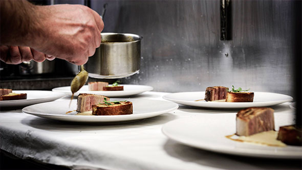 Serving process: chef&rsquo;s hand pours dark sauce from a metal ladle over portions of roasted meat or steaks, served on minimalist white plates in a row