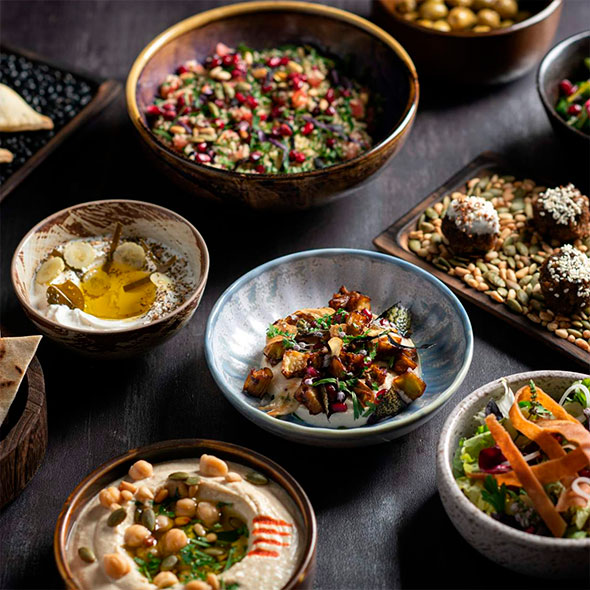 Assortment of traditional Lebanese meze appetizers (hummus, salads, pita, pomegranate, and olive snacks) in various bowls on a dark table.
