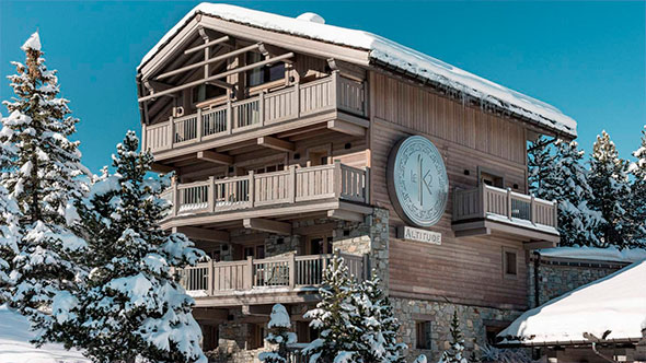 Winter exterior of one of the Le K2 Altitude chalets, made of wood and stone, featuring balconies, a snow-covered roof, and a large circular hotel logo.