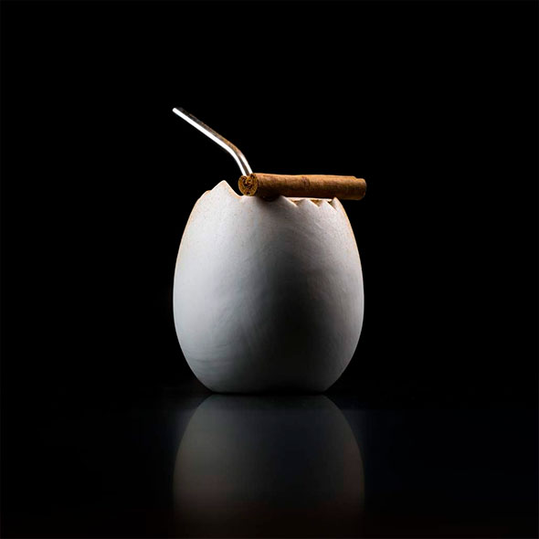 Signature cocktail or drink from Le K2 Palace, presented in a stylized white bowl resembling an eggshell, with a straw and a thin cigar resting on top, against a dark background.