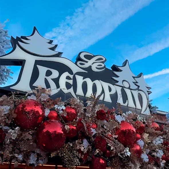 Sign of Le Tremplin bar: close-up in winter season, where white stylized letters and a logo with fir trees are placed above ample decor of red disco balls (mirror balls), gold and white branches and flowers.