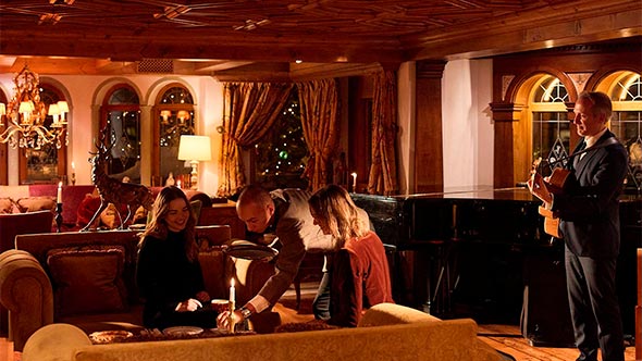 Cozy lounge of the Bar / Tea Room Airelles: guests seated on luxurious sofas with velvet-upholstered cushions, in the center of an interior decorated with wood panels and a carved ceiling; a grand piano and a singer in a suit with a guitar are visible on the right.