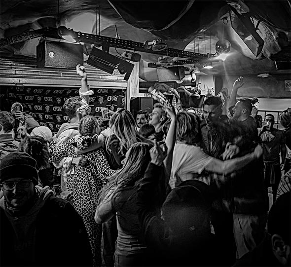 Black and white photo of a lively party at Prends ta Luge: a dense crowd of people, some in ski hats, dancing and hugging, with hands raised, beneath a low ceiling with visible speakers.