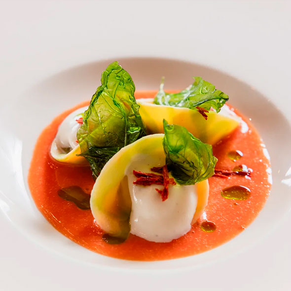 Exquisite pasta or ravioli dish, served in tomato sauce with cheese balls and green leaves on a pristine white plate.