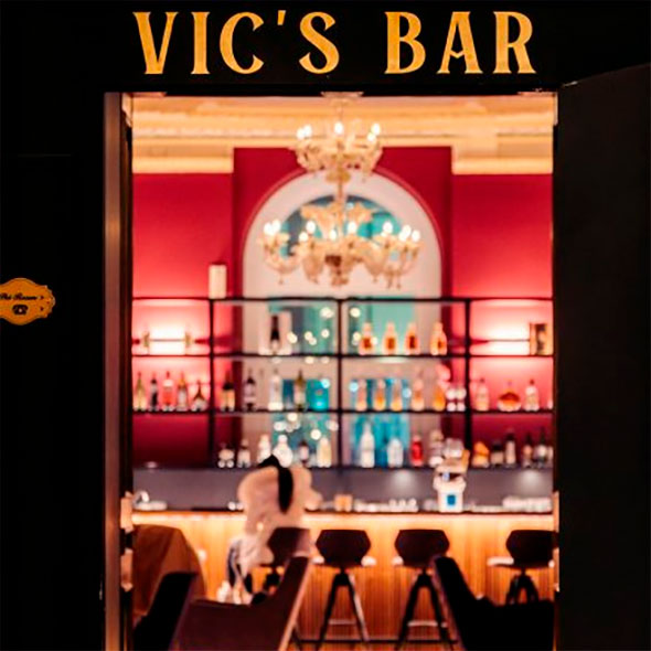 Entrance to Vic&rsquo;s Bar with a gold sign, revealing an interior with red walls, an illuminated bar counter, and a chandelier.